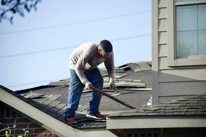 A roofer tearing off damaged asphalt shingles beside a dormer.